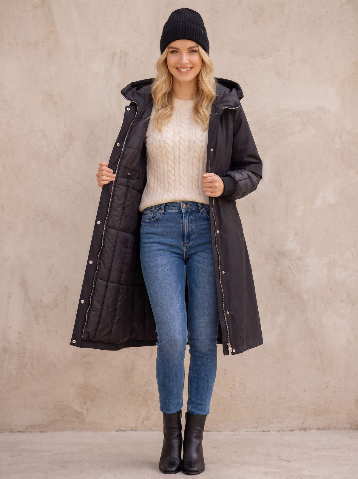 Дълго черно яке парка Benny Boutique с капитонирани ръкави и качулка, носено от усмихната руса жена с плетена шапка и дънки.