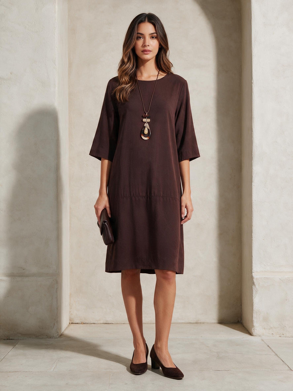 Кафява рокля Benny Boutique с ниска талия и ръкави три четвърти, представена върху модел с дълго колие с висулка.