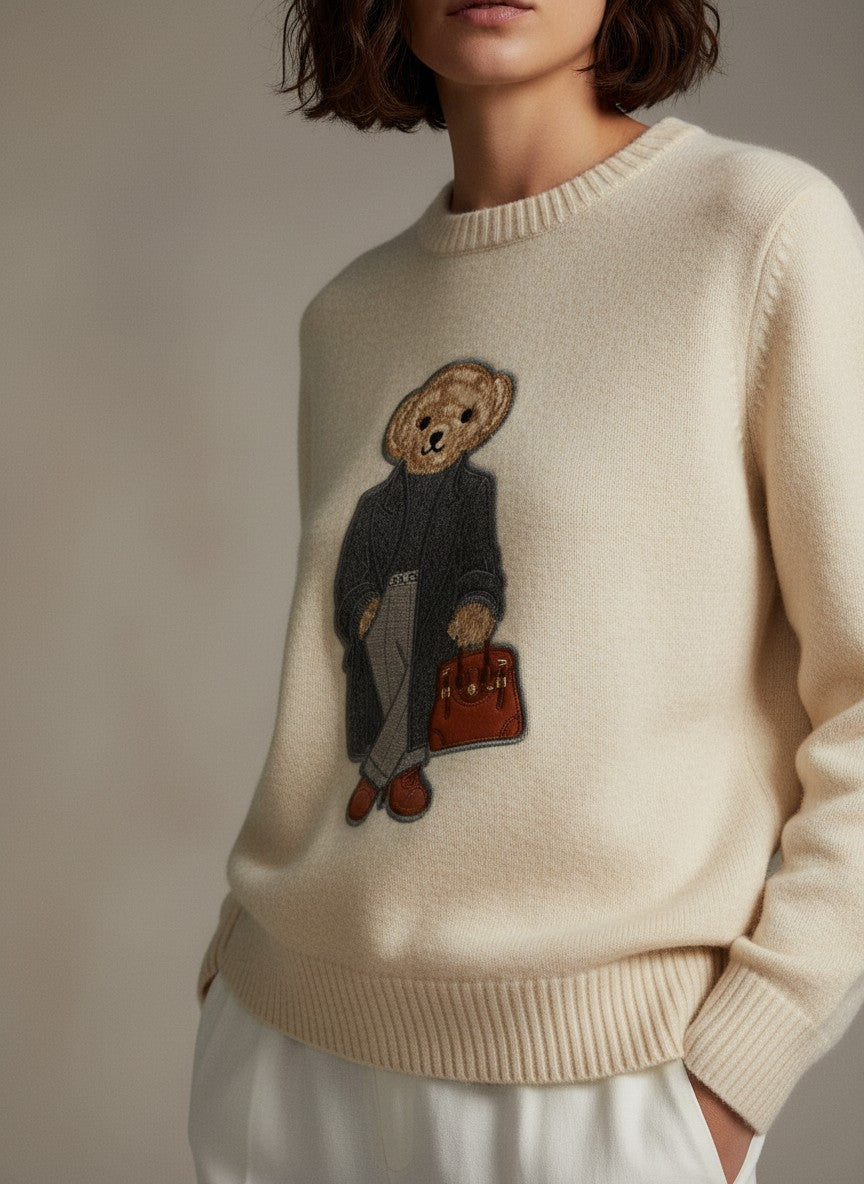 Кремав финоплетен пуловер Benny Boutique с игрива апликация на мече, облечено с палто и държащо чанта.