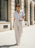 Benny Boutique бяла късръкавна риза с черни точки и предно връзване, носена с бежови панталони на градска улица.