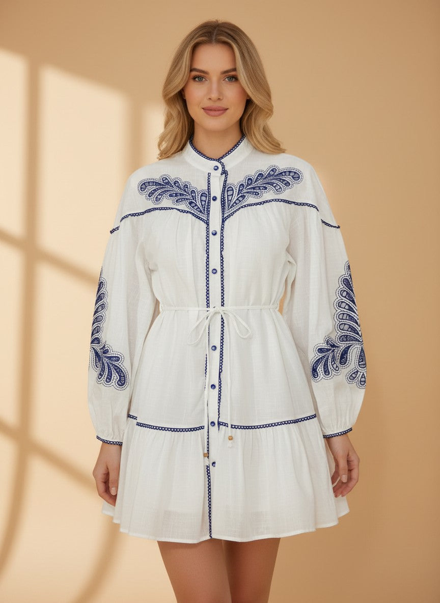 Бяла мини рокля Benny Boutique със синя бродерия, дълги ръкави и талия с връзки, представена върху русокос модел.