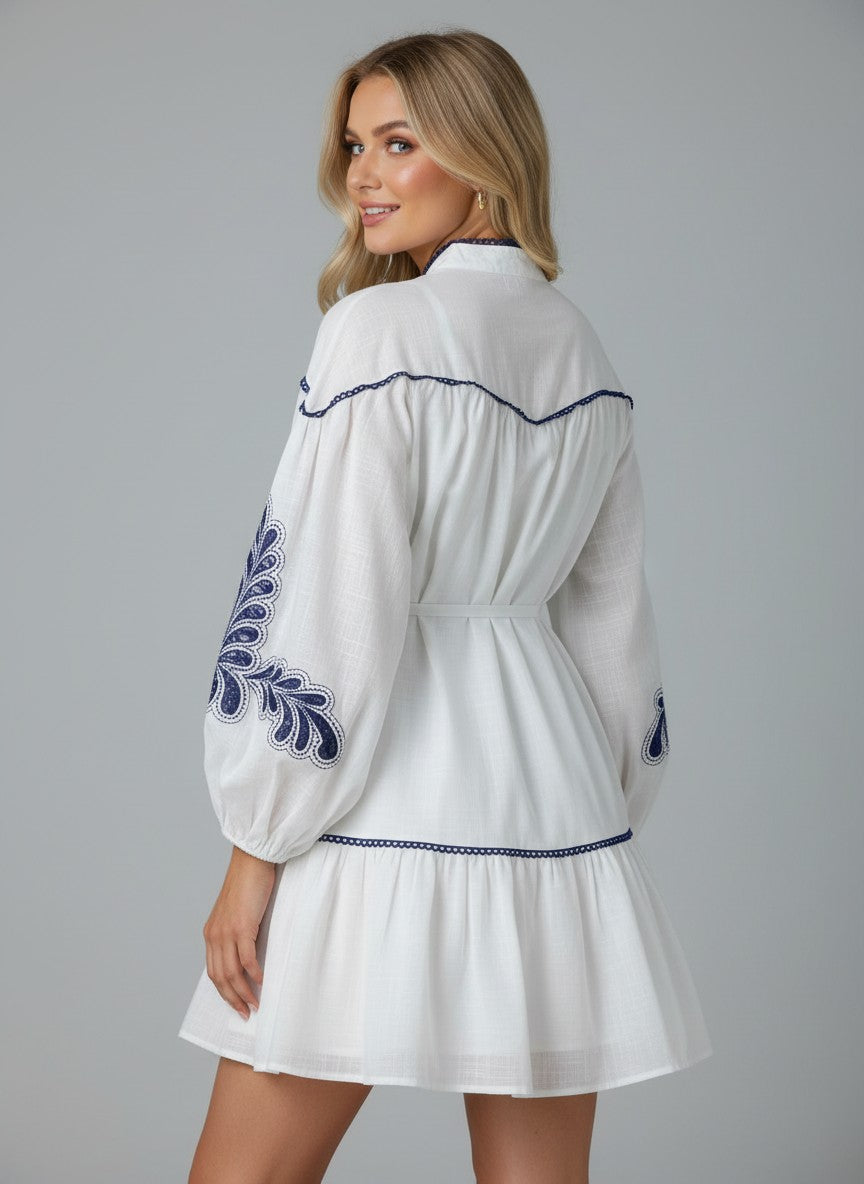 Бяла мини рокля Benny Boutique със синя бродерия, дълги ръкави и връзки на талията, представена върху рус модел.